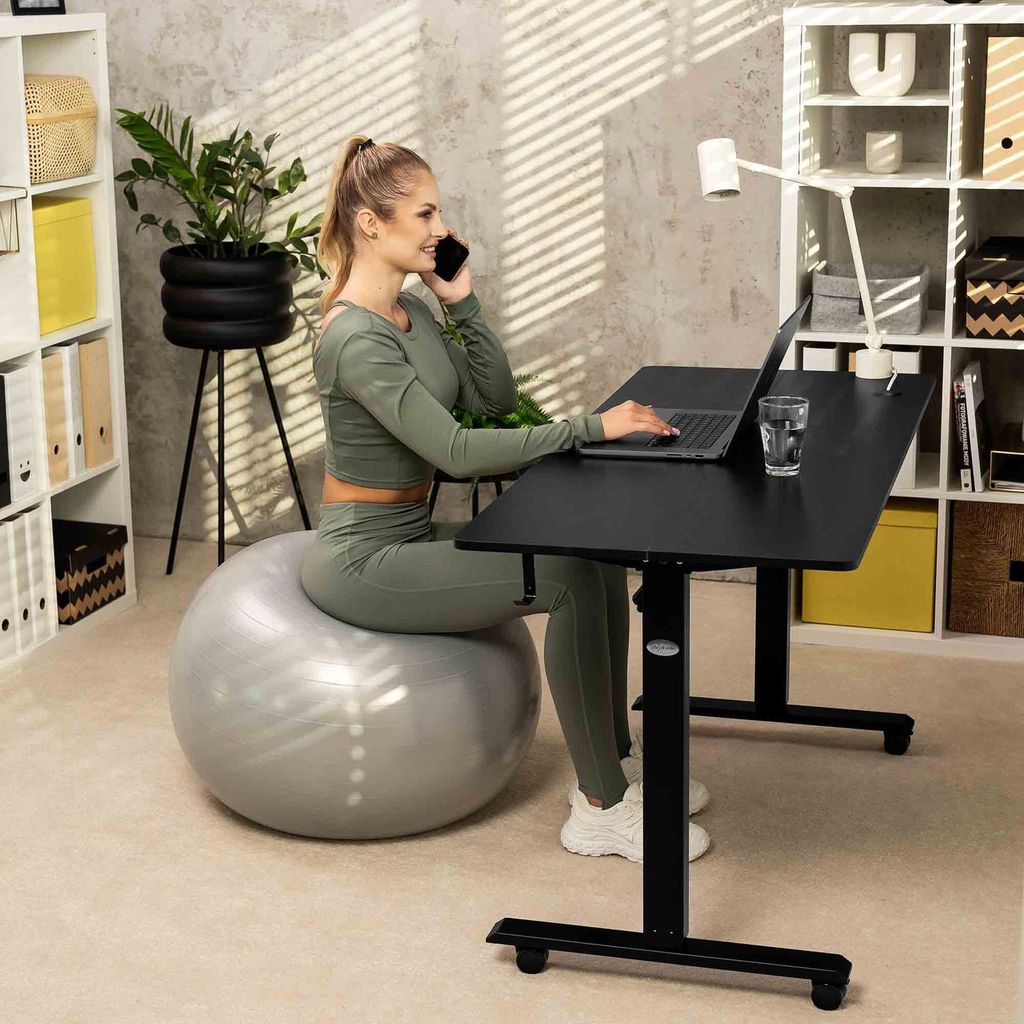 A woman is working on a laptop while sitting on an exercise ball at a standing desk in a home office.