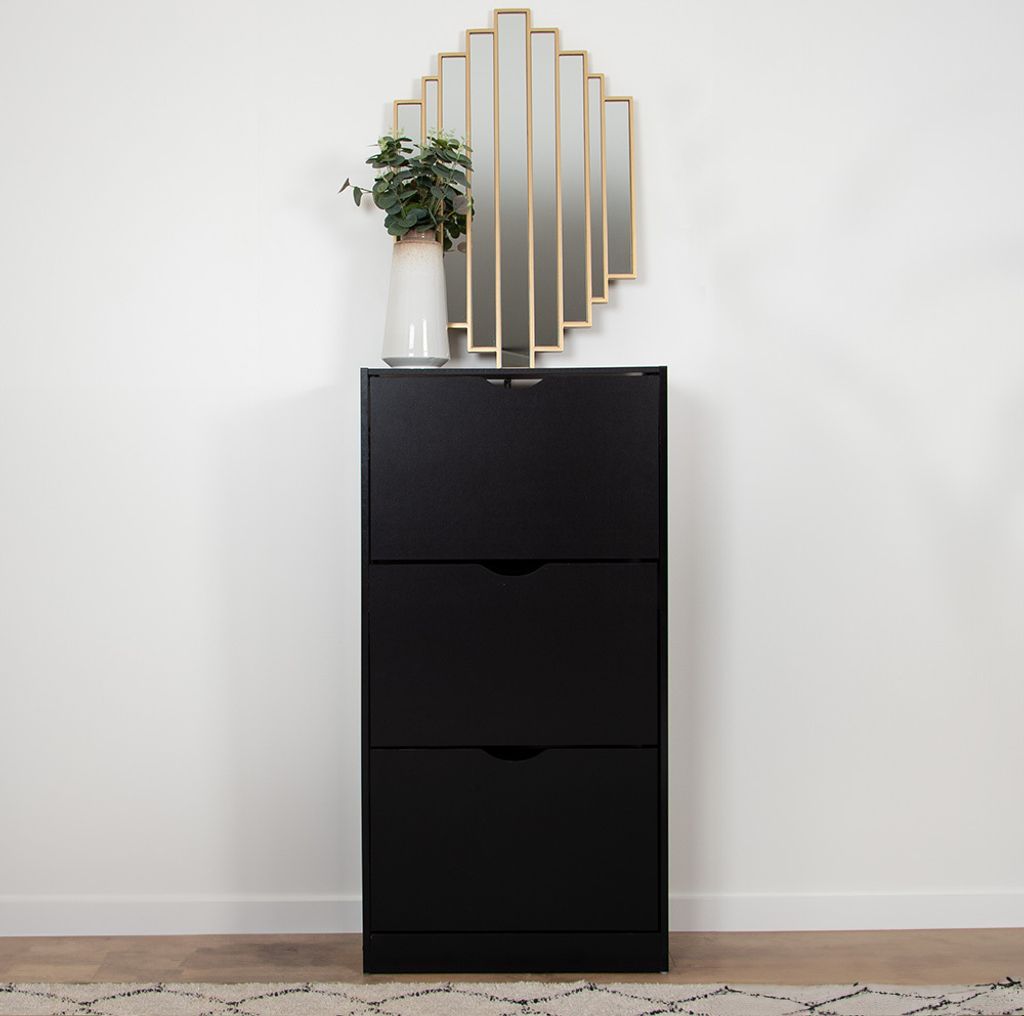 A black shoe cabinet with three drawers against a white wall, topped with a decorative mirror and a vase with a plant.