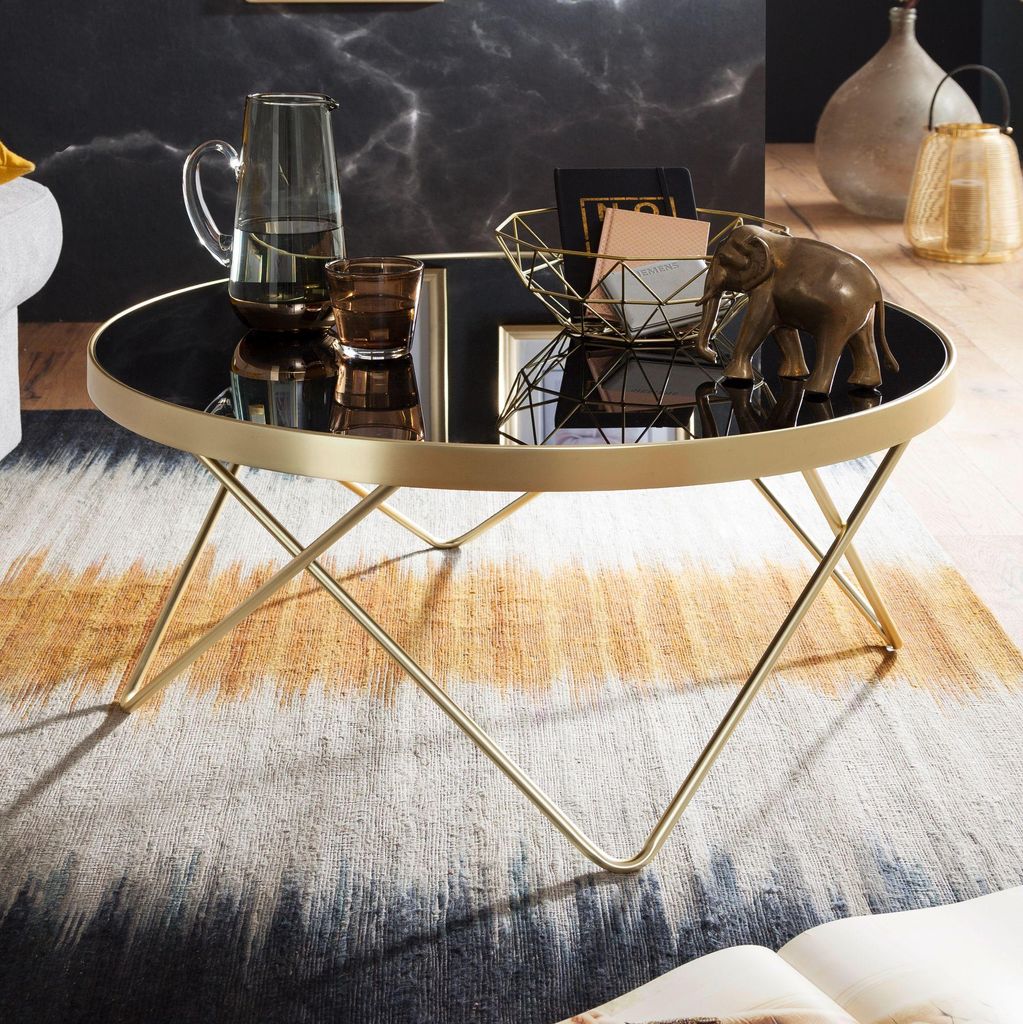A modern coffee table with a black glass top and gold metal legs, set on a patterned rug in a living room.