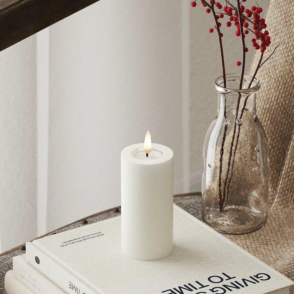 A white LED candle with a lit flame sits on top of a stack of books, next to a glass vase with red berries.