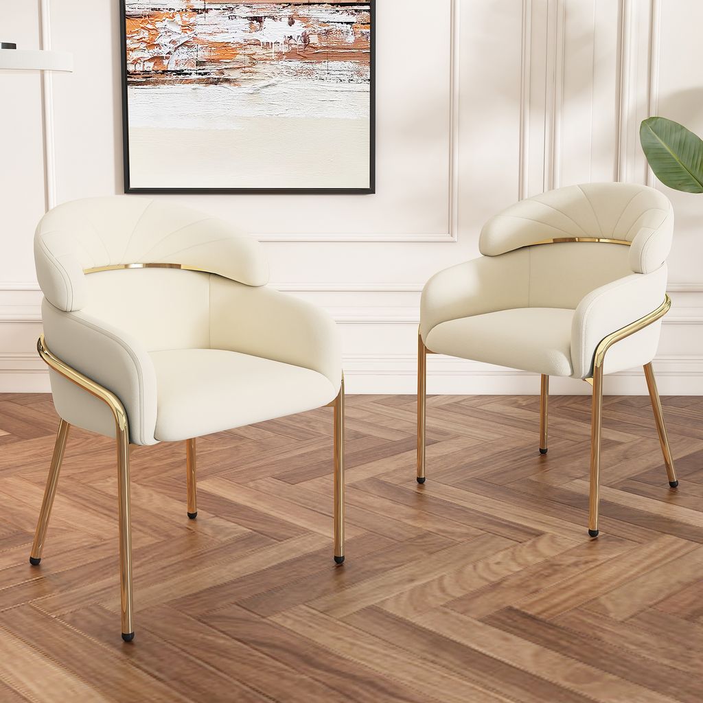 Two beige armchairs with gold legs are placed on a wooden floor in a room with white paneled walls and a painting on the wall.