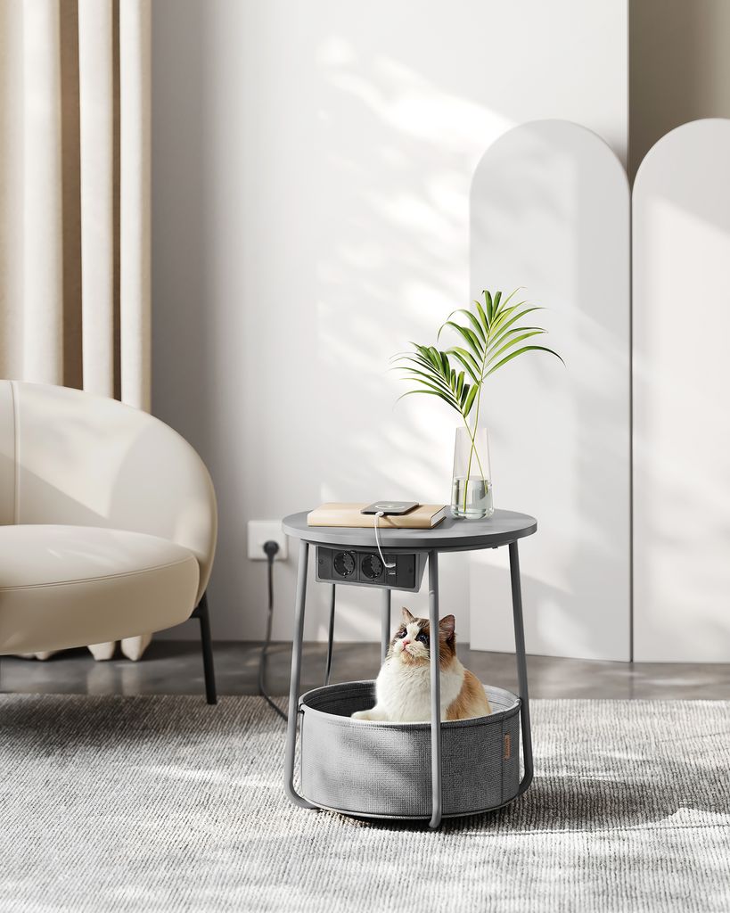 A cat is sitting in a pet bed integrated into a round side table with built-in power outlets and USB ports. The table has a plant in a vase on top, and a phone is charging. A chair is visible in the background.