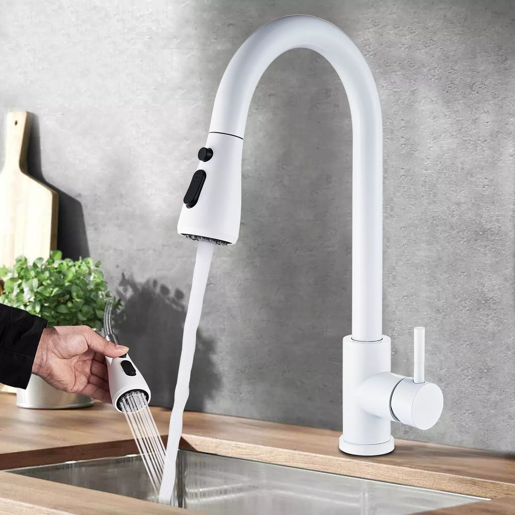 A white kitchen faucet with a pull-out spray head is shown in use over a stainless steel sink. A hand is holding the spray head, demonstrating its functionality.