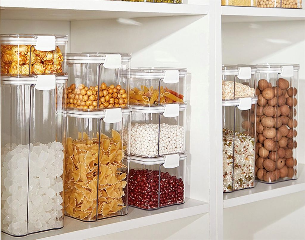 A collection of clear plastic food storage containers filled with various dry goods, neatly stacked on white shelves.