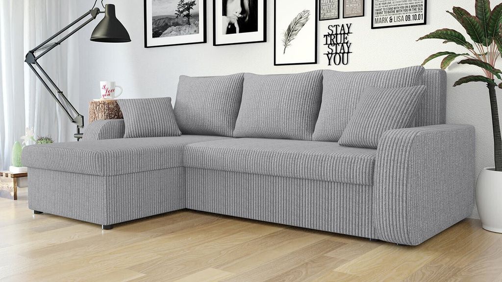 A gray sectional sofa with pillows in a living room setting, with framed pictures and a floor lamp in the background.