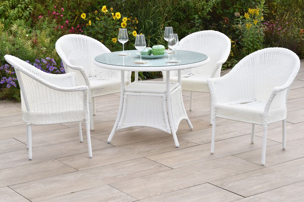 A white wicker patio set with a glass-topped table and four chairs is set up on a stone patio, surrounded by garden flowers.