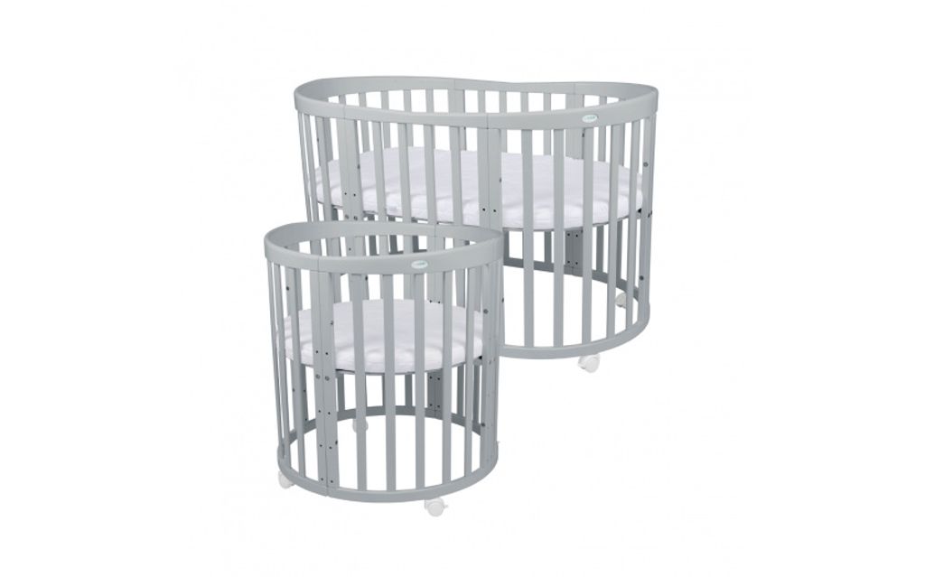 Two baby cribs of different sizes, one larger and oval-shaped, the other smaller and cylindrical, both are gray with white mattresses and are on wheels.