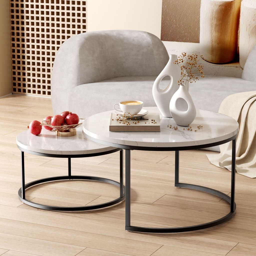 A set of two round nesting coffee tables with white marble-like tops and black metal bases are placed on a light wood floor. One table holds a bowl of red apples, and the other has a coffee cup and two decorative vases. A neutral-colored sofa is in the bac