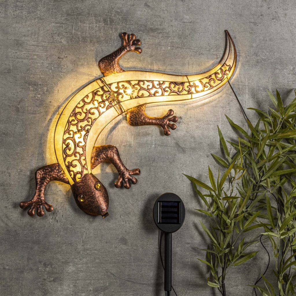 A decorative solar-powered gecko wall light is mounted on a textured gray wall, with a small solar panel stake and some greenery visible.