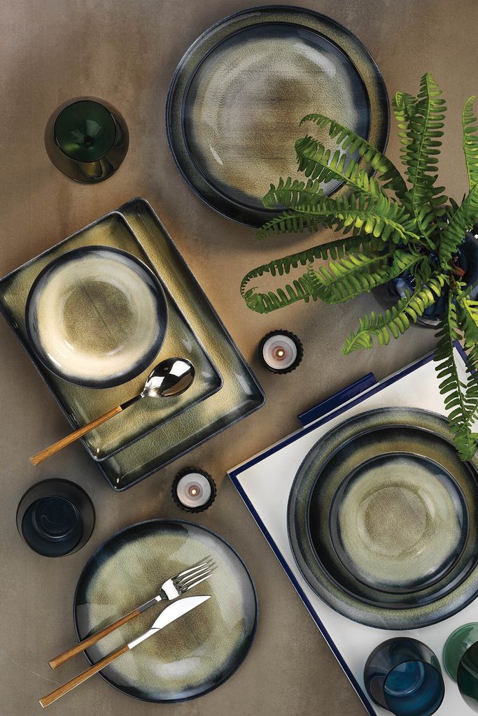 A table setting with dark blue and beige ceramic plates, bowls, cutlery, and a glass, arranged with a fern plant and candles.