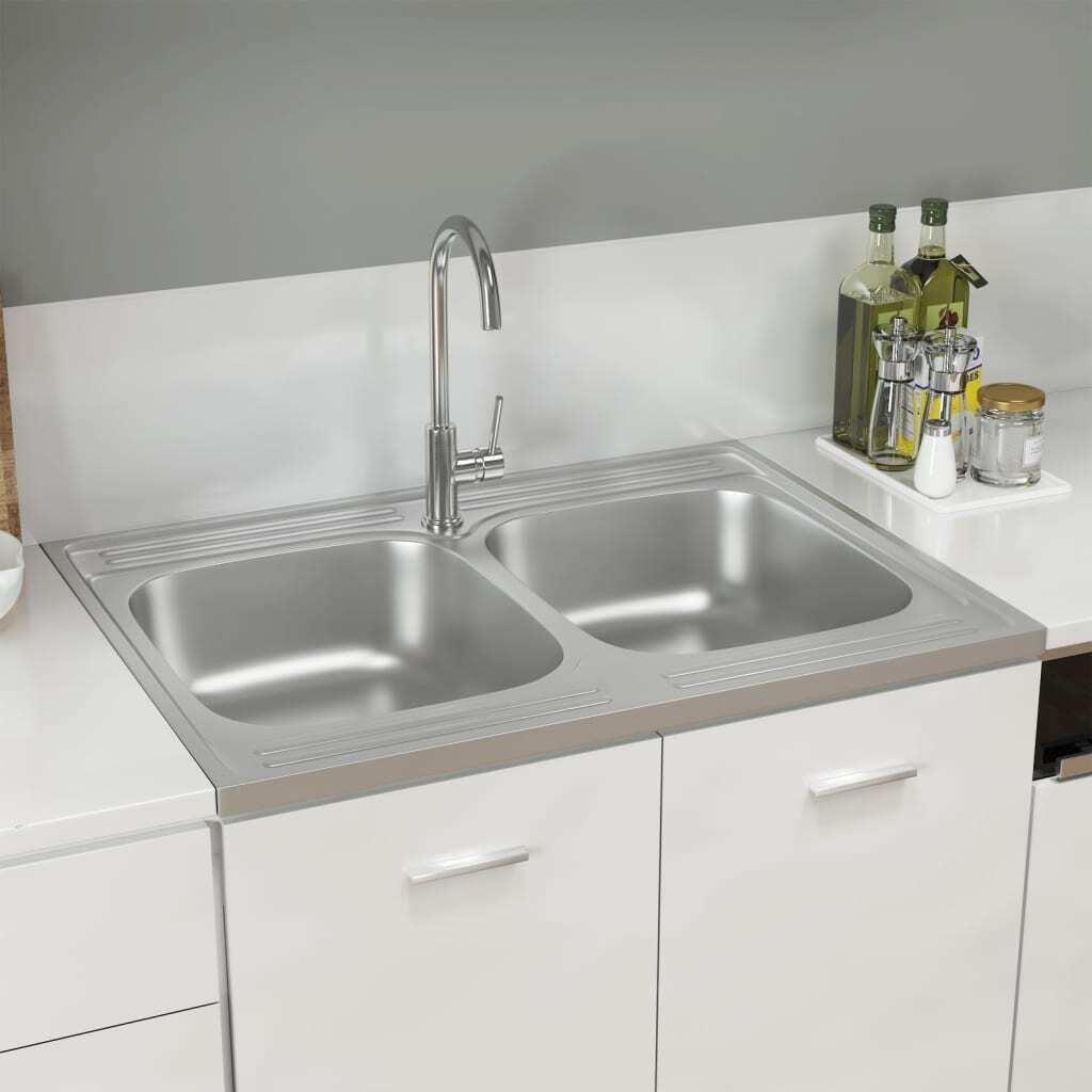 A stainless steel double sink with a faucet, set into a white countertop and cabinets, with some kitchen items in the background.
