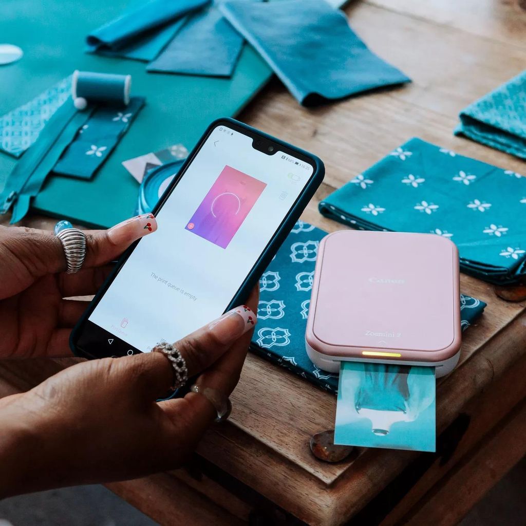 A person is holding a smartphone and a pink Canon Zoemini 2 photo printer is printing a photo.