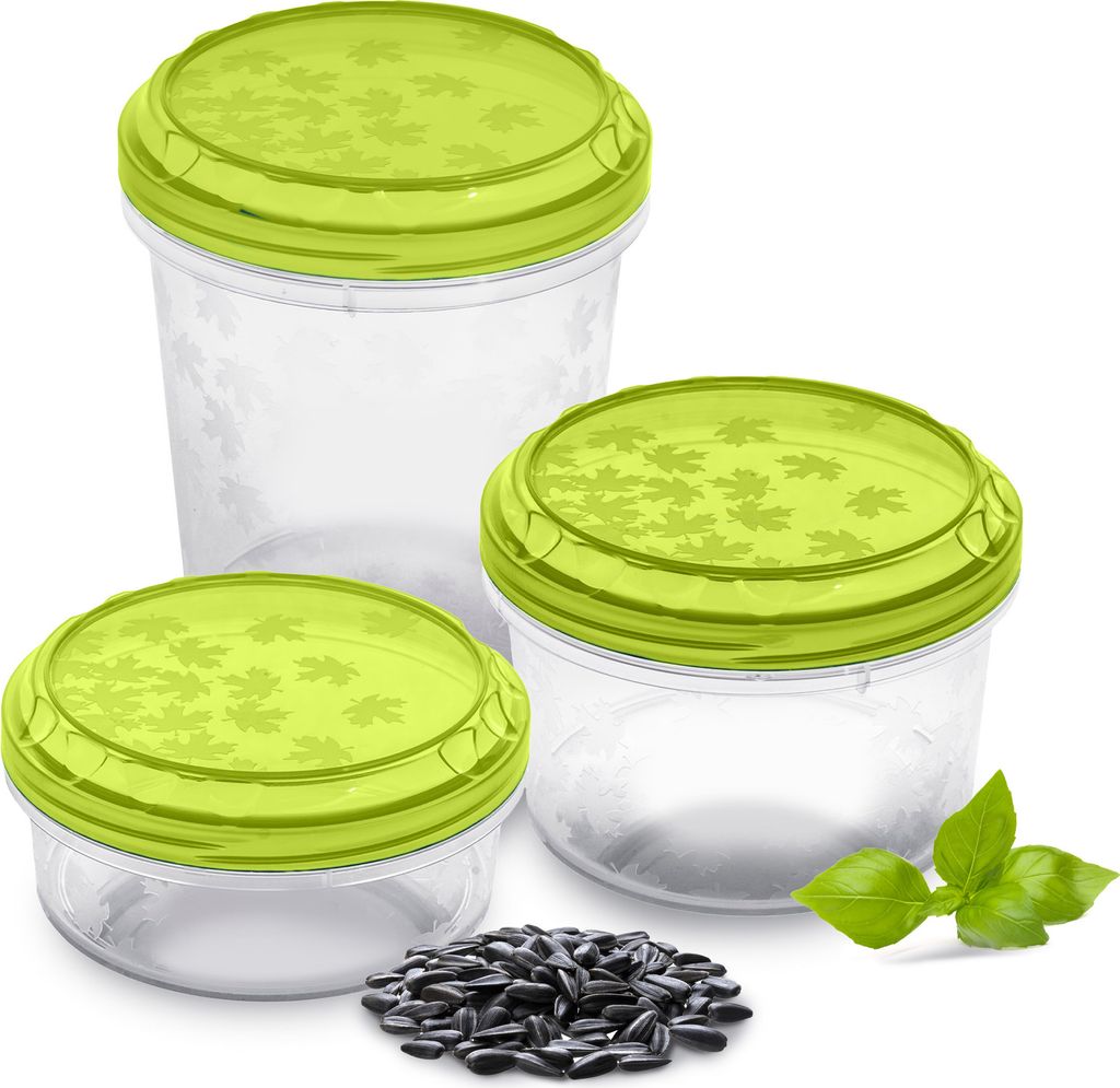 Three clear plastic food storage containers with green lids are arranged on a white background, with a small pile of sunflower seeds and a sprig of basil in the foreground.