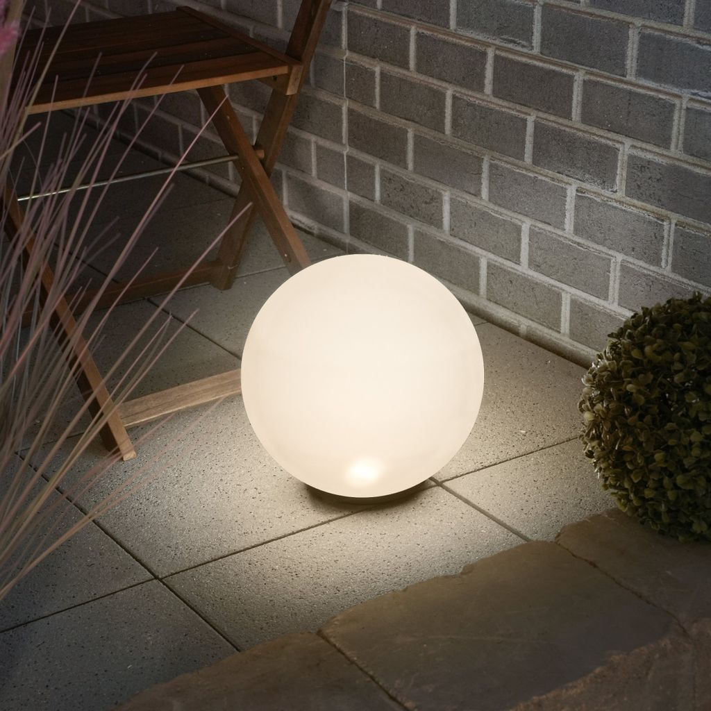 A glowing sphere-shaped lamp sits on a tiled patio next to a wooden chair and some dry grass.