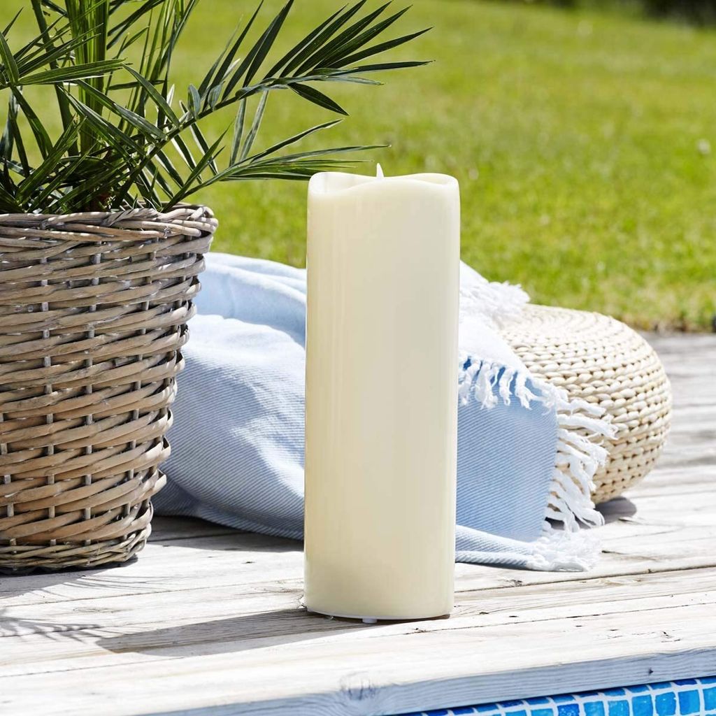 A large LED candle sits on a wooden deck next to a potted plant and a blue blanket.