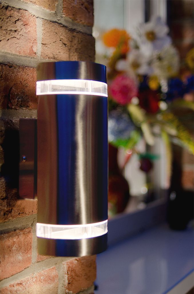 A modern, cylindrical outdoor wall light mounted on a brick wall, with a blurred background of flowers.