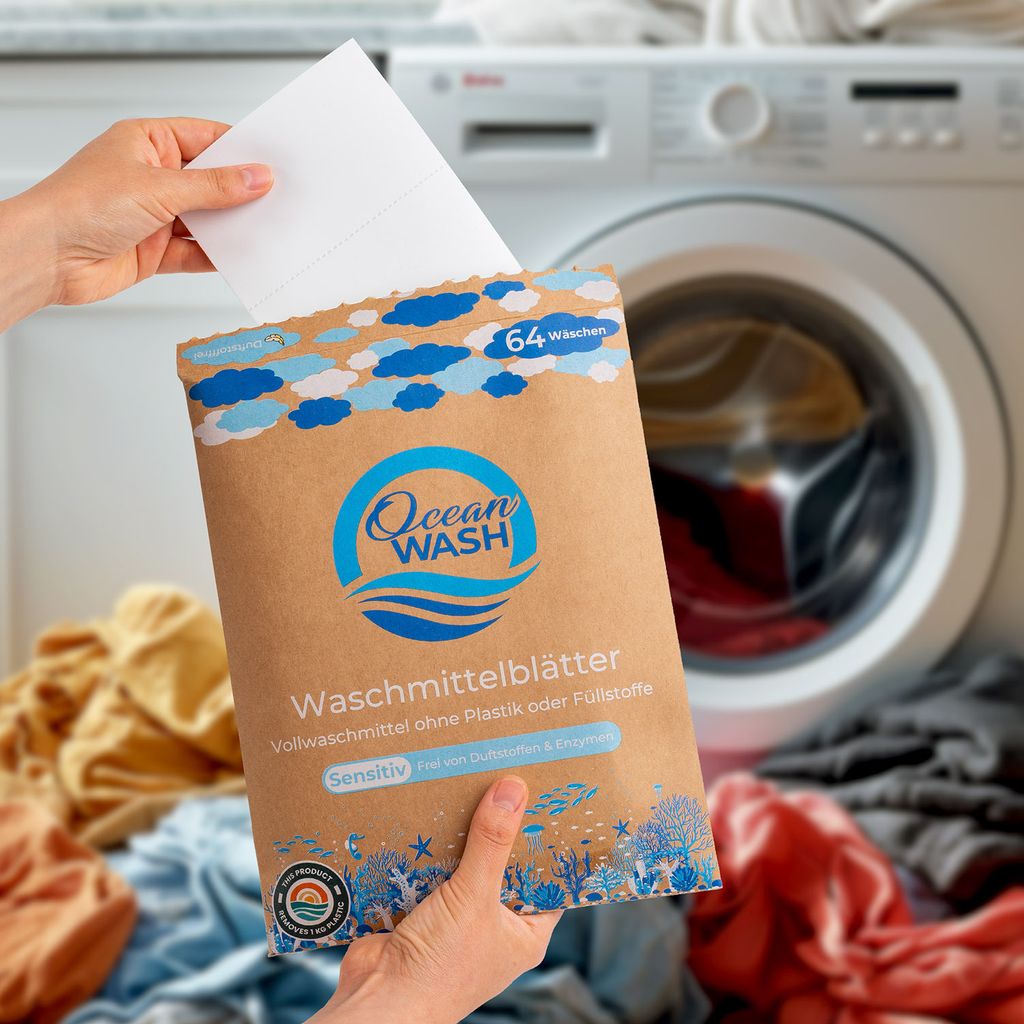 A person is holding a package of OceanWash laundry detergent sheets in front of a washing machine with laundry around it.