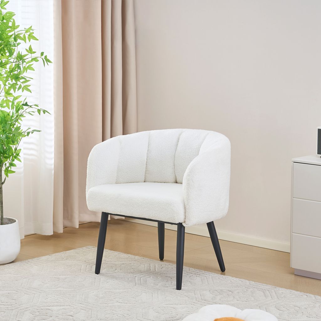 A white fluffy armchair with black legs sits on a light-colored rug in a room with beige curtains and a plant.
