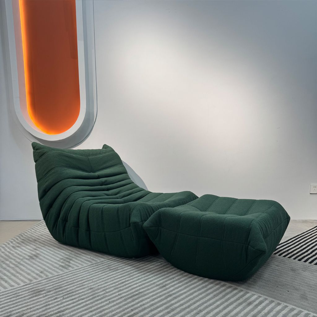 A green sofa and ottoman are placed on a striped rug in a room with a white wall and an orange accent light.
