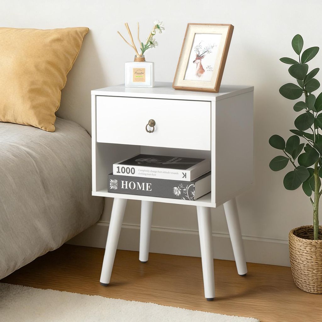 A white nightstand with a drawer and an open shelf, placed next to a bed. Decorative items are on top of the nightstand.