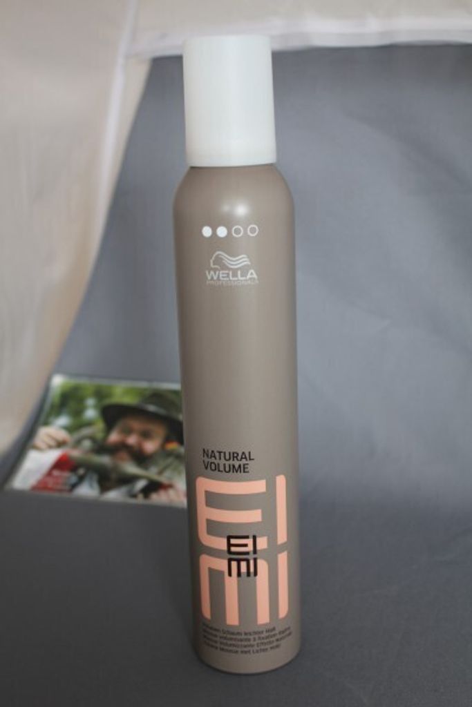 A bottle of Wella EIMI Natural Volume Styling Mousse is shown in the foreground, with a blurred image of a person in the background.