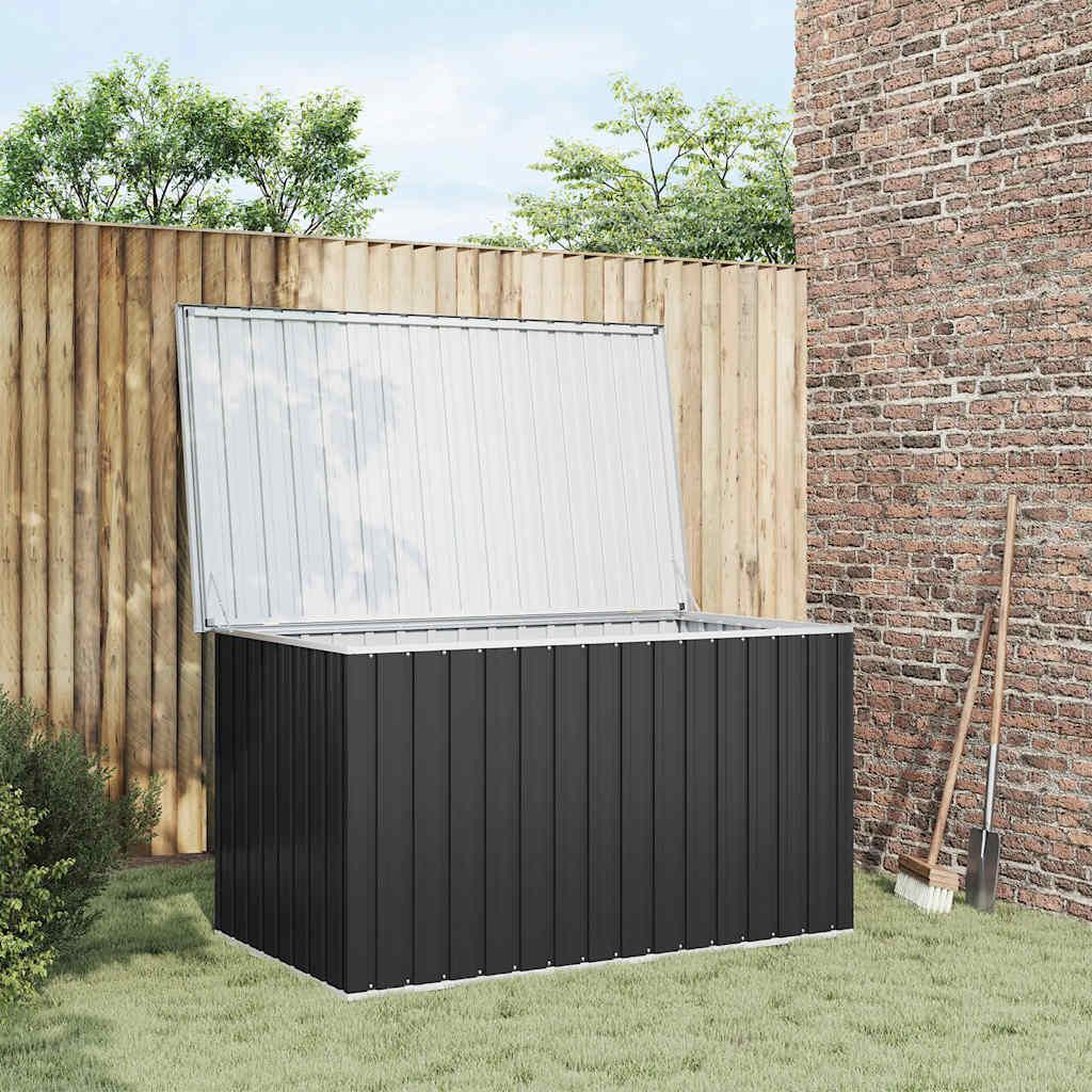 An anthracite garden box with its lid open, placed on a grassy lawn next to a wooden fence and a brick wall. Gardening tools are leaning against the wall.