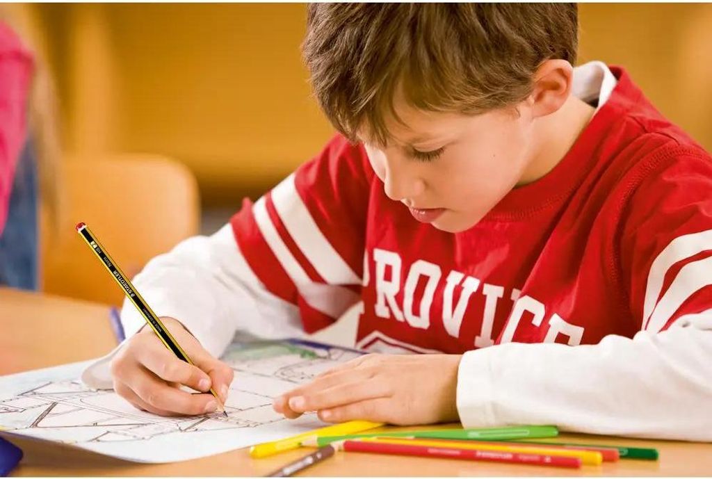 A young boy is drawing with a pencil on a piece of paper, with colored pencils in front of him.