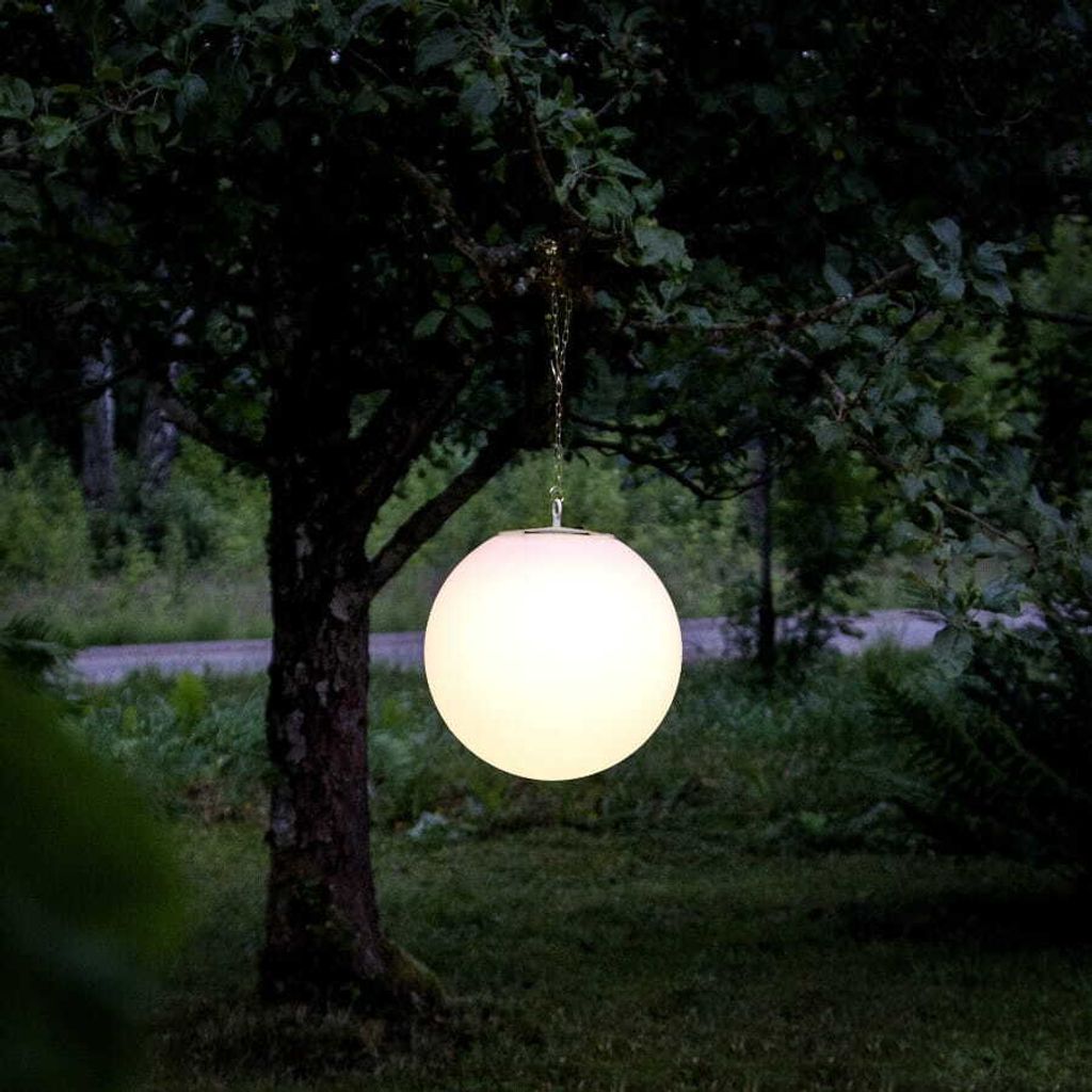 A glowing white sphere hangs from a chain attached to a tree branch in a garden at dusk.