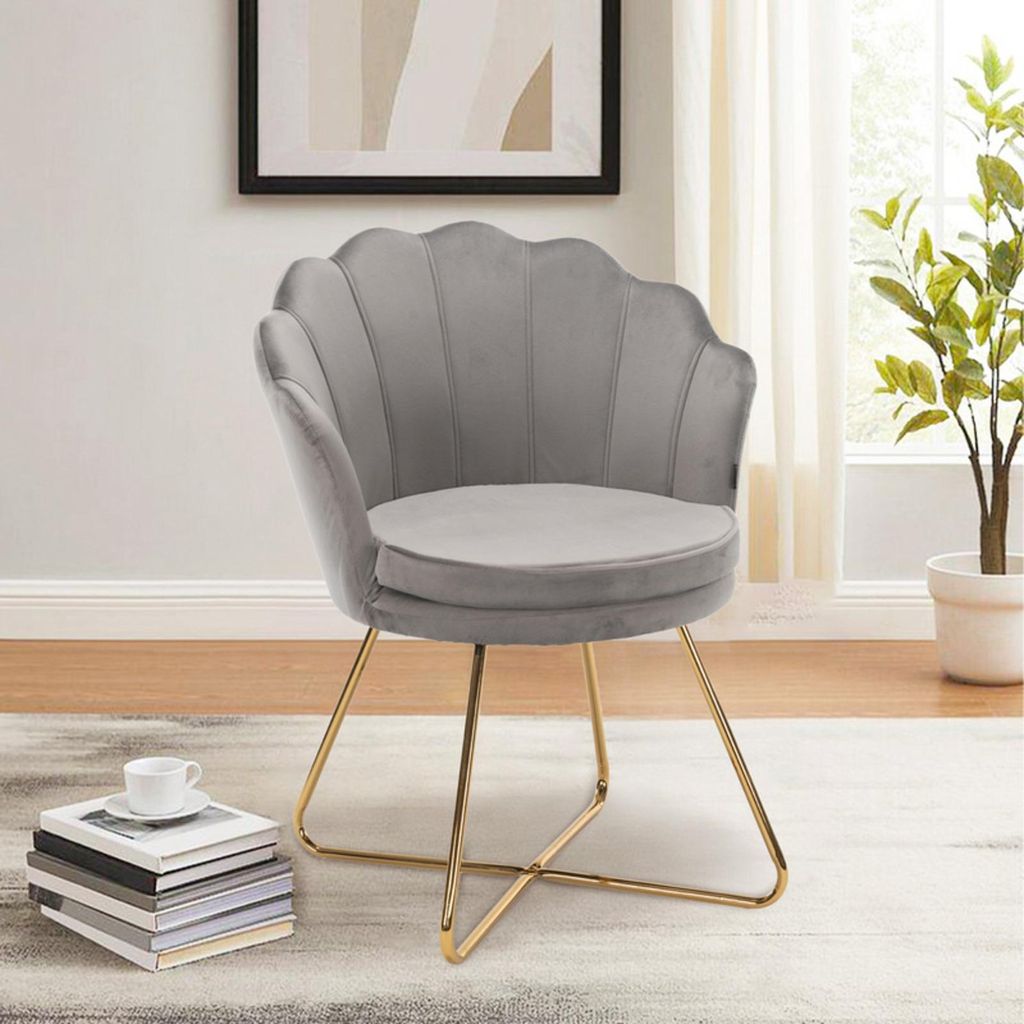 A gray velvet accent chair with gold metal legs is shown in a well-lit room with a plant and books in the foreground.