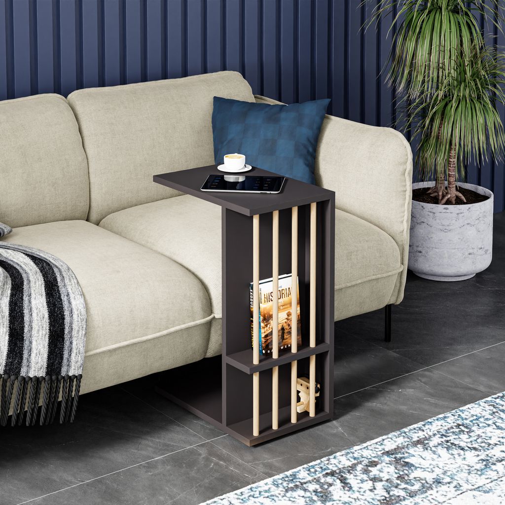 A dark gray side table with vertical wooden slats is placed next to a beige sofa in a living room setting. A tablet and a cup of coffee are on the table, and a book is in the lower shelf. A blanket is draped over the sofa, and a potted plant is visible in 