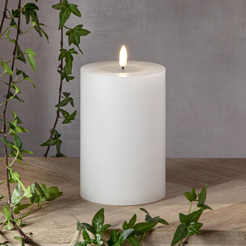 A white LED candle with a flame sits on a wooden surface, with ivy vines framing the scene against a grey background.