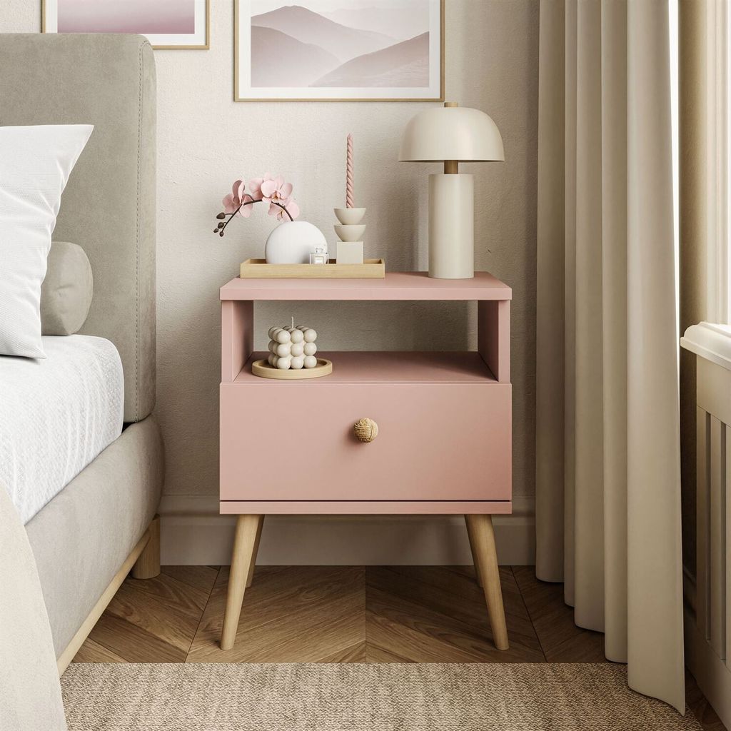A pink nightstand with a drawer and an open shelf, decorated with a lamp, candle, and a small decorative item.