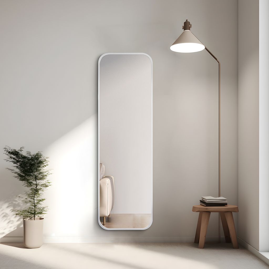 A full-length mirror is placed against a wall next to a potted plant and a floor lamp. A small wooden stool with folded towels sits beside the mirror.