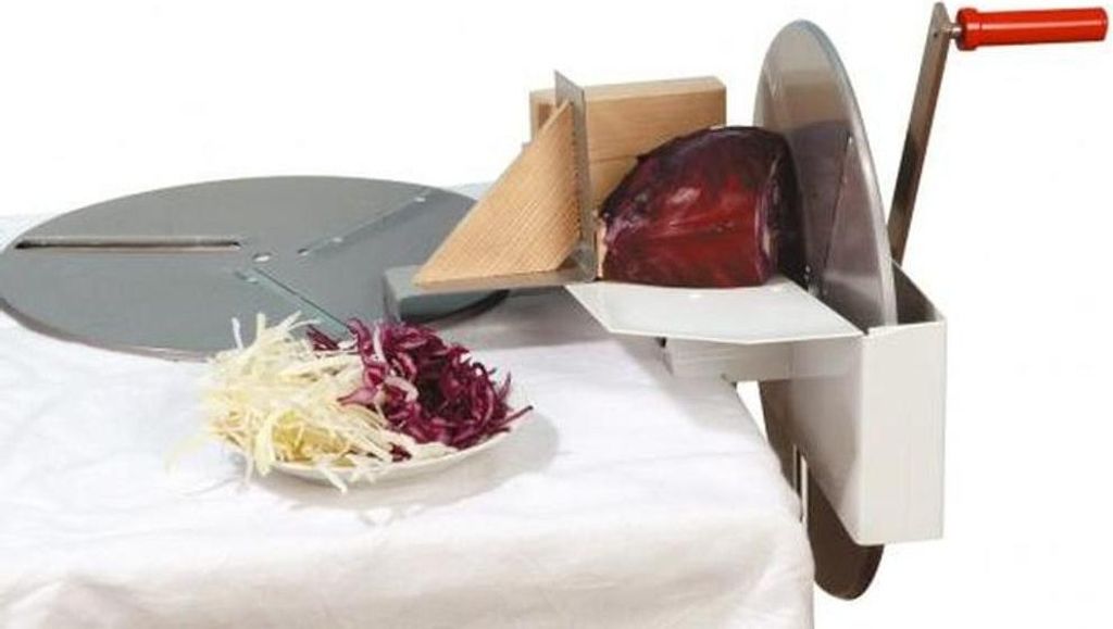 A red cabbage is being sliced by a manual slicer, with shredded cabbage on a plate in the foreground.