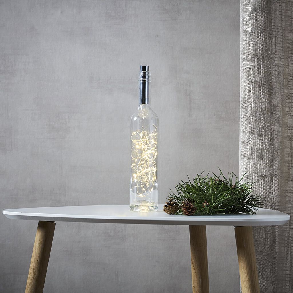 A glass bottle filled with warm white LED lights sits on a white table next to a sprig of pine and pinecones.