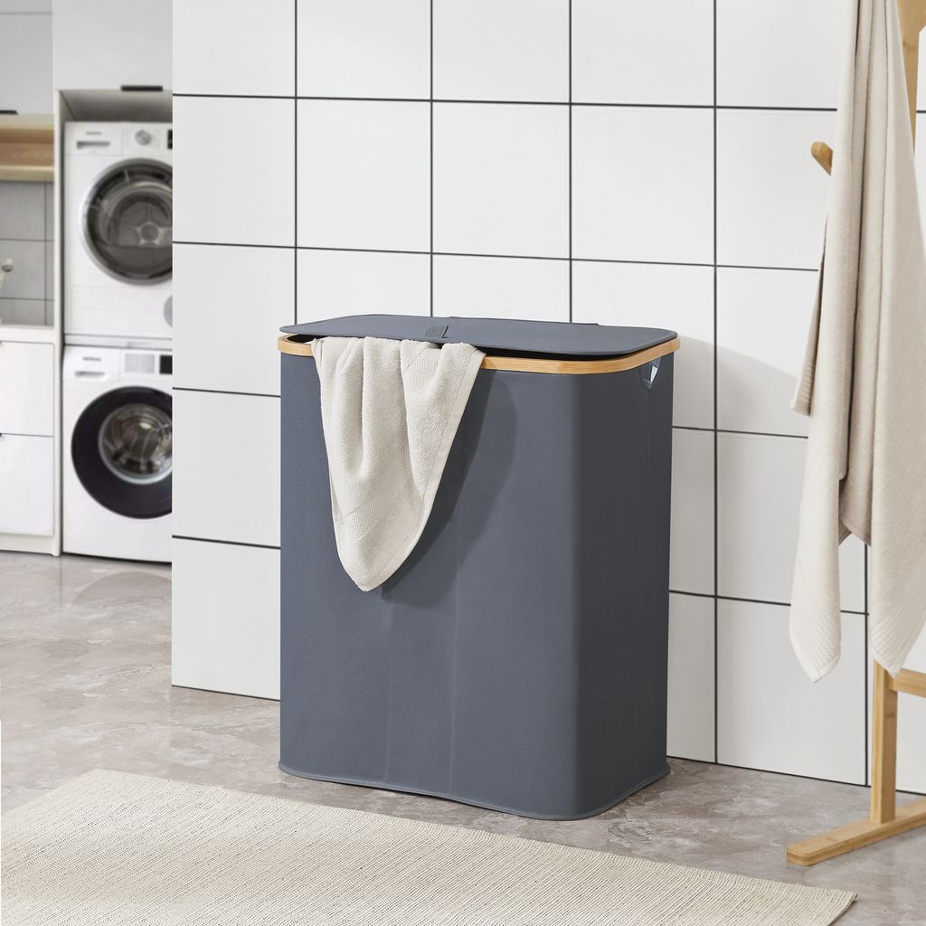 A dark gray laundry basket with a beige towel draped over it, placed in a laundry room with washing machines and tiled walls.