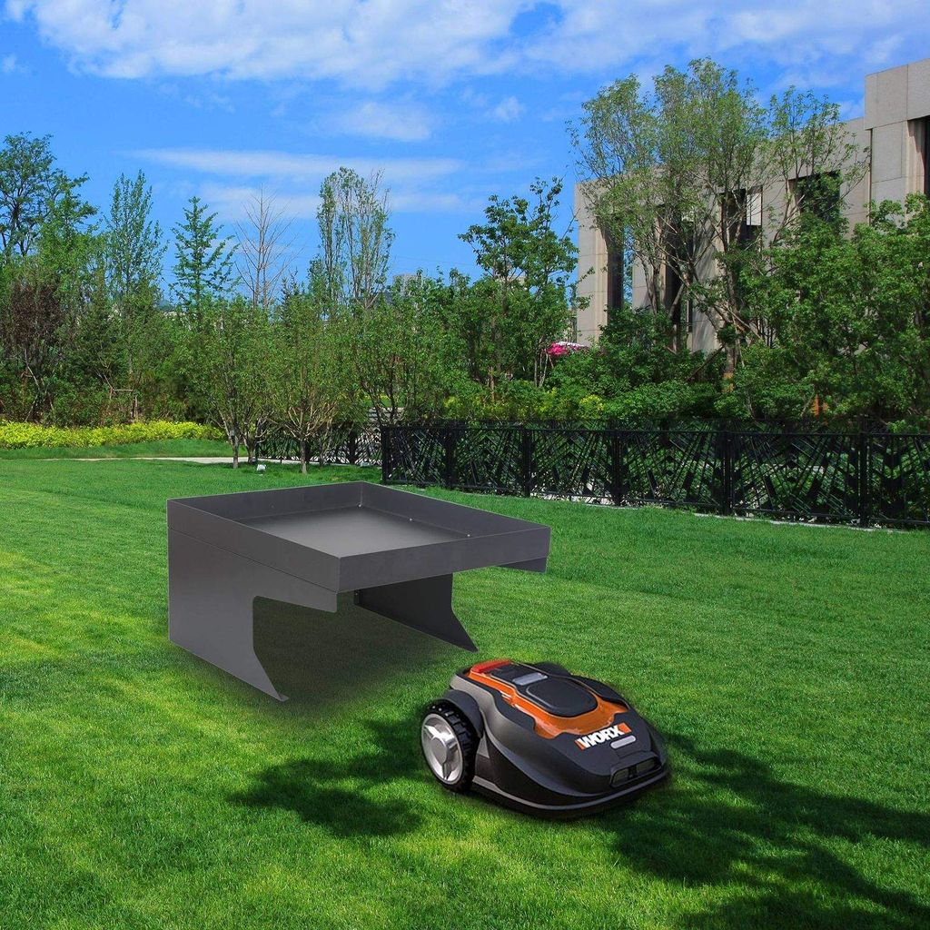 A robotic lawnmower is parked next to a protective garage structure on a green lawn, with trees and a modern building in the background.