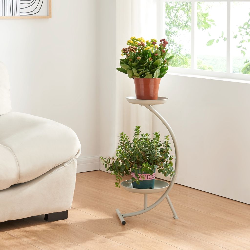 A two-tiered plant stand with two potted plants is placed next to a sofa in a well-lit room with a window.
