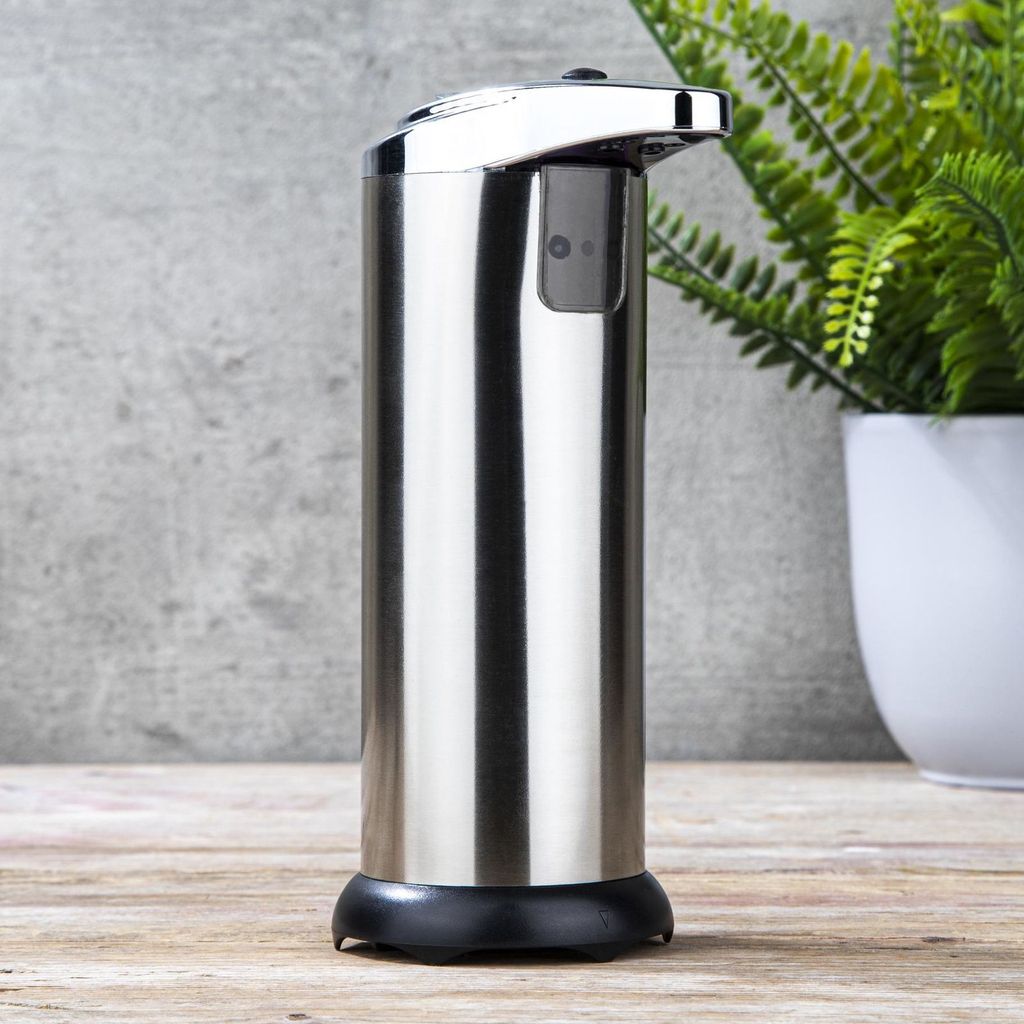 A stainless steel automatic soap and disinfectant dispenser is shown in a studio setting with a concrete wall and a potted plant in the background.