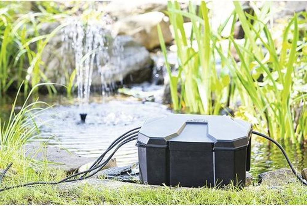 A black and gray HEISSNER Box for a garden socket is placed on the grass next to a pond with a fountain.