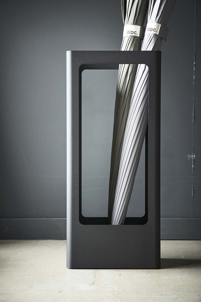 A modern, minimalist umbrella stand with umbrellas inside, set against a dark gray wall and a light gray floor.