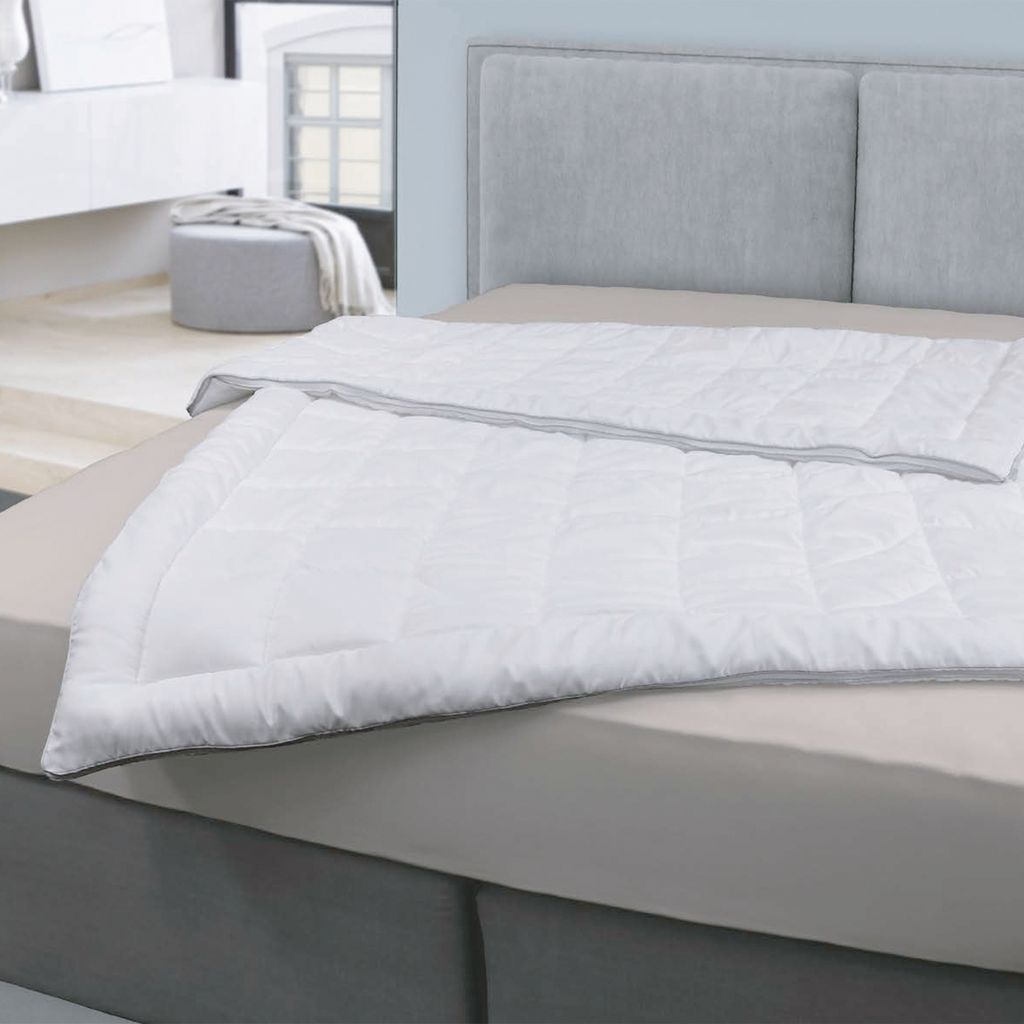 A white duvet is laid out on a bed with a grey headboard in the background. A grey ottoman and a dresser are visible in the background.