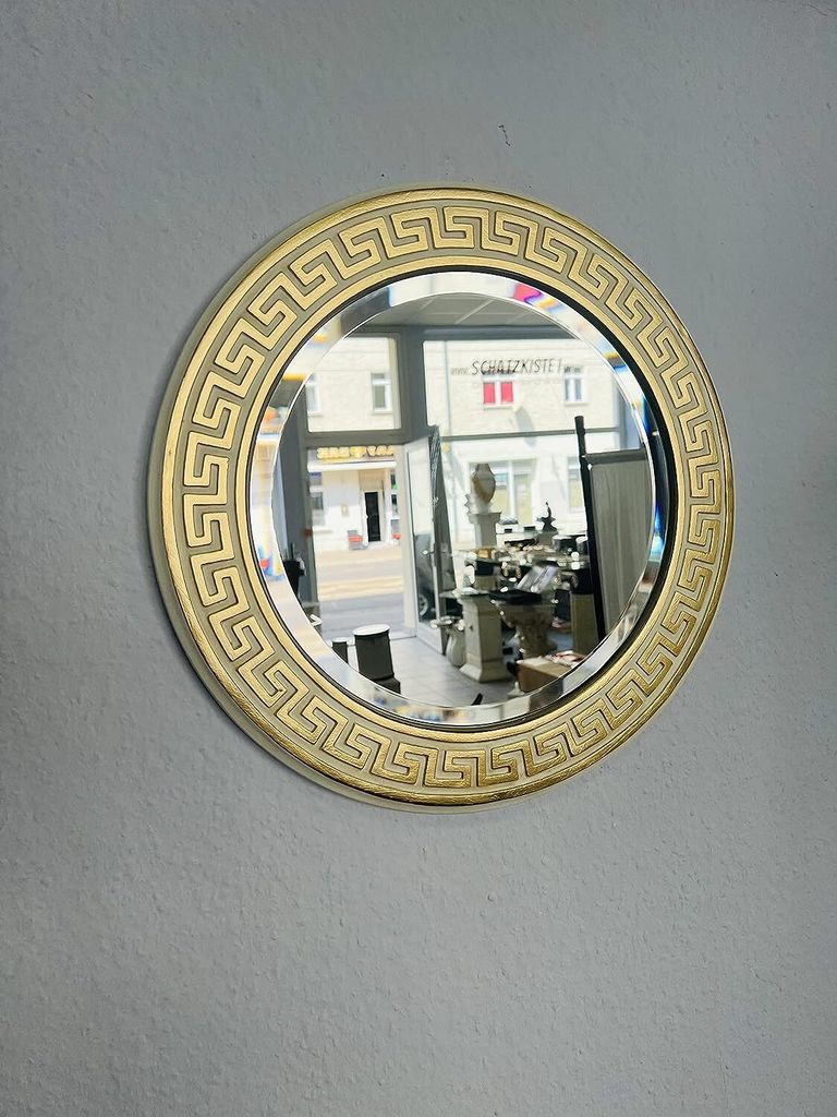 A round mirror with a gold ornate frame hangs on a light gray wall. The mirror reflects a shop interior.