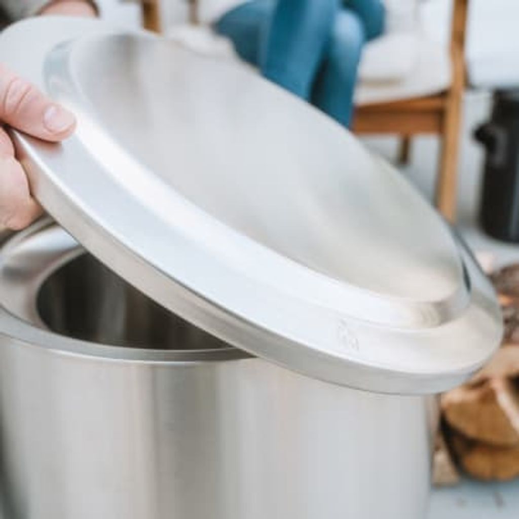 A hand is lifting the lid off a stainless steel fire pit. In the background, people are sitting on chairs and there is a stack of firewood.
