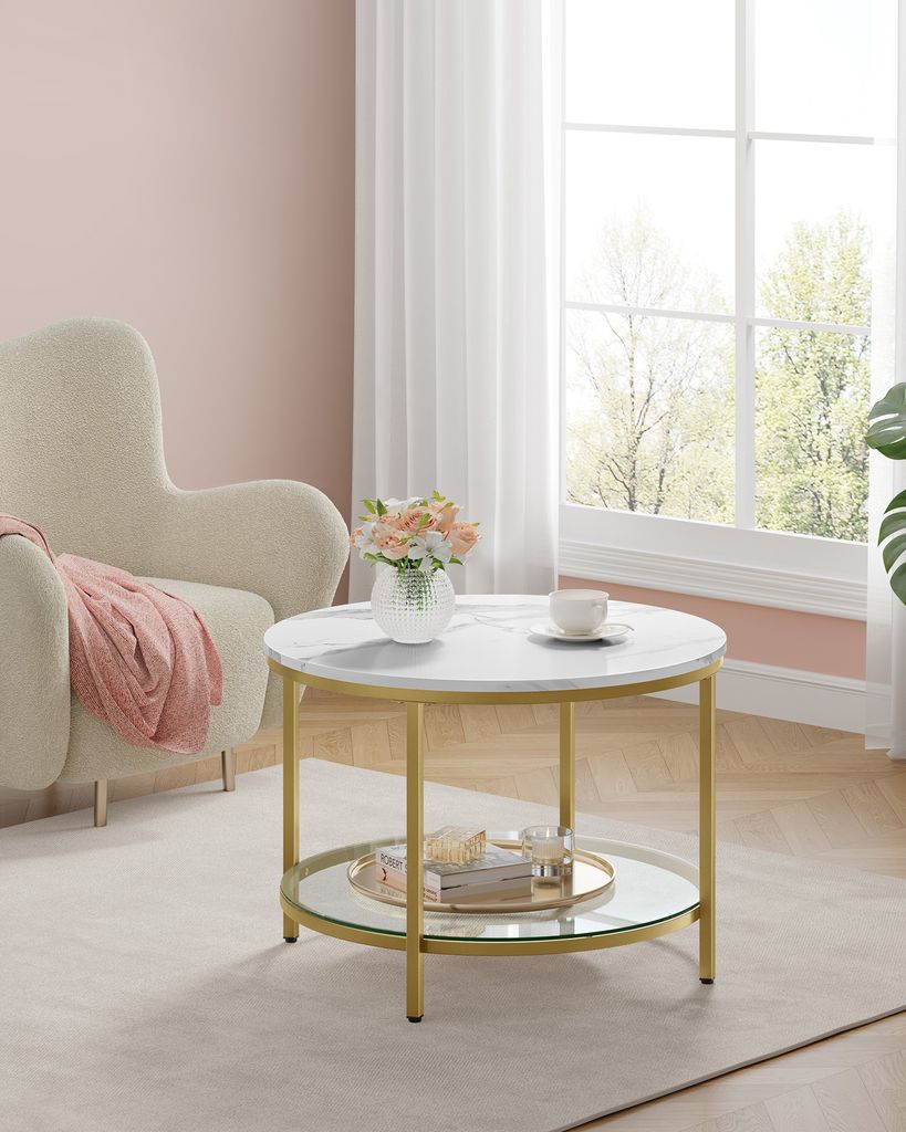 A round coffee table with a marble top and gold legs, placed in a living room setting next to an armchair.