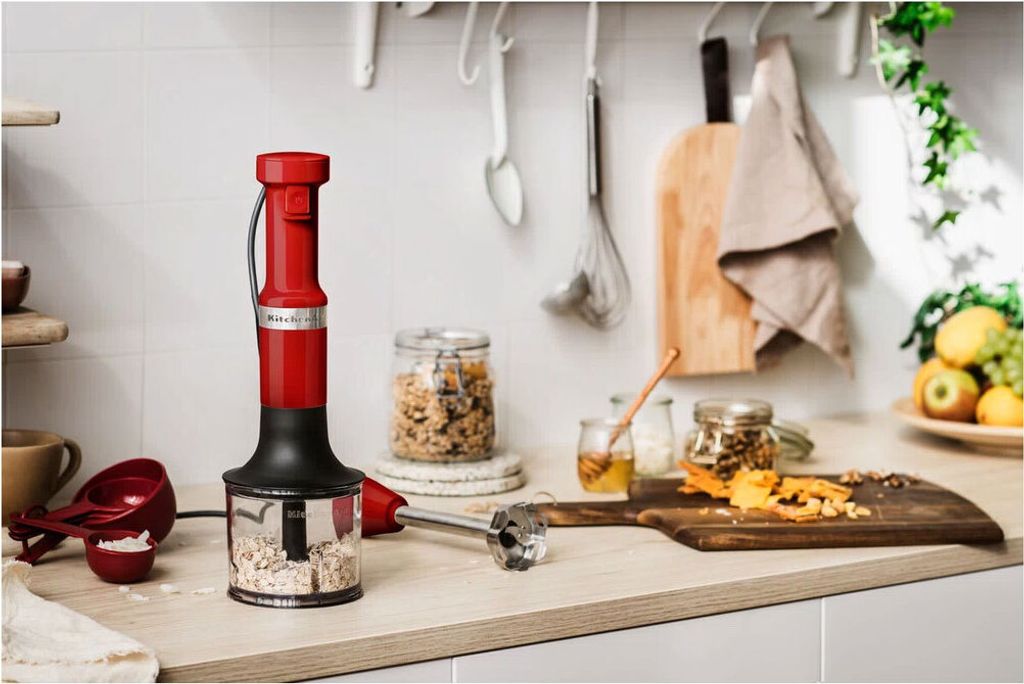A red KitchenAid immersion blender is shown on a kitchen counter with ingredients and kitchen utensils in the background.