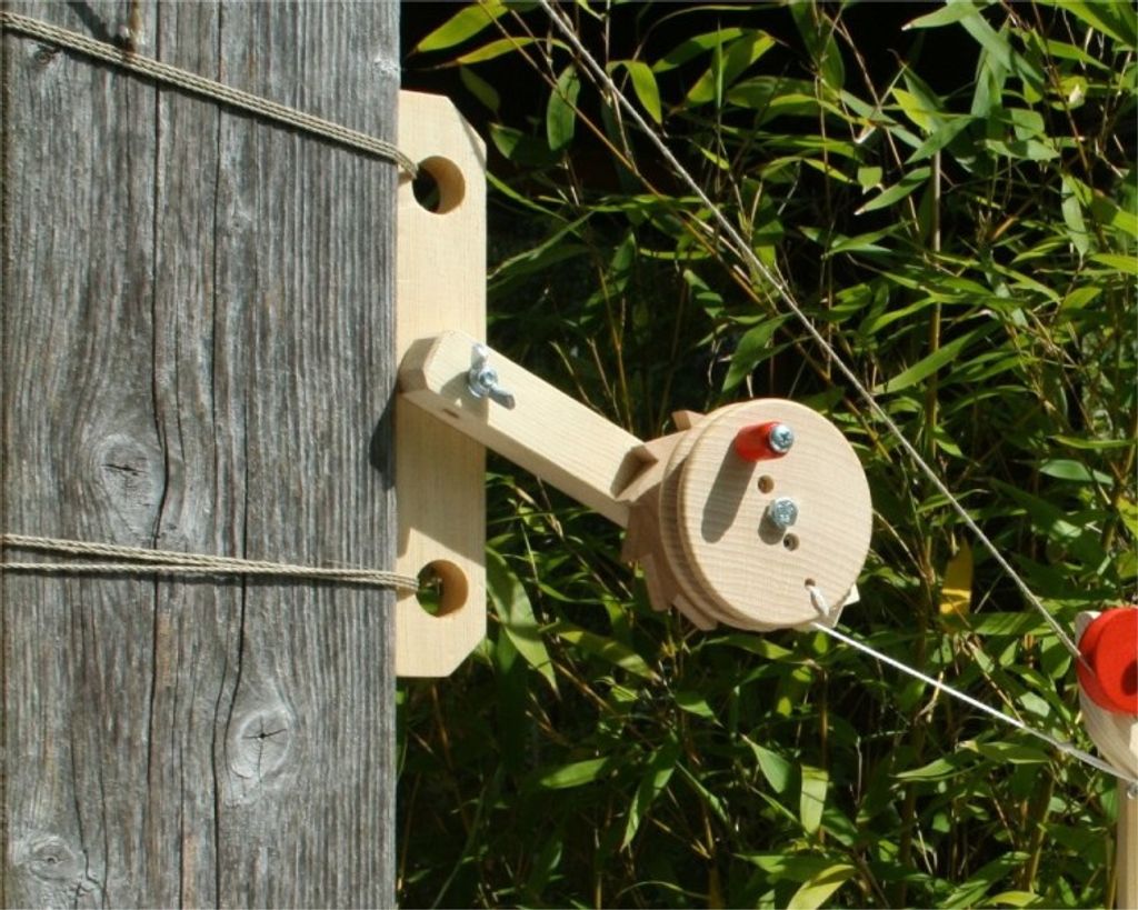 A wooden winch mechanism attached to a wooden post, with green foliage in the background.