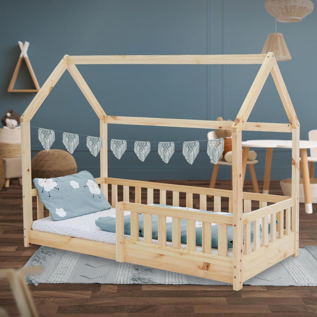 A wooden house-shaped bed for children, set up in a room with a blue wall and a rug on the floor.