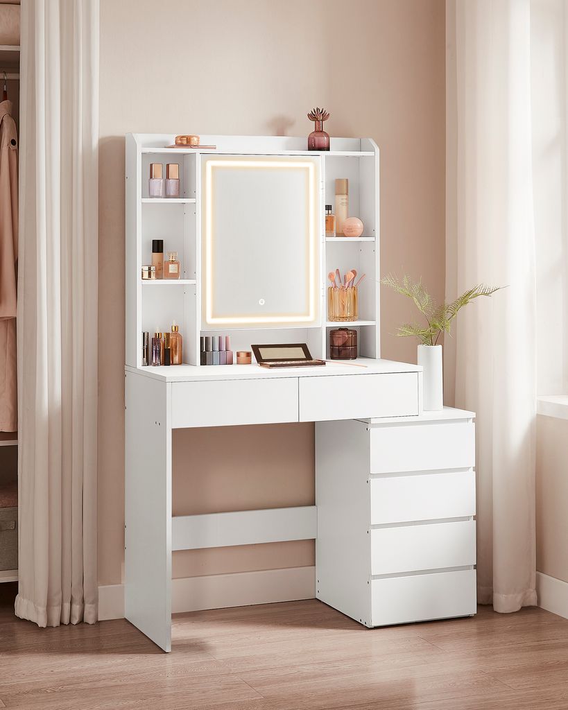 A white vanity table with a lighted mirror and drawers, set against a light pink wall and curtains.