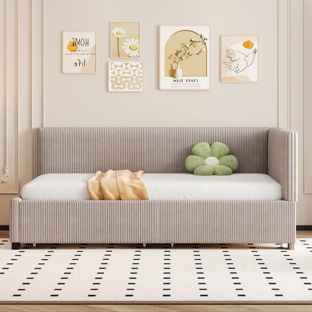 A daybed with a mattress, a yellow blanket, and a green flower-shaped pillow is shown in a room with artwork on the wall and a patterned rug on the floor.